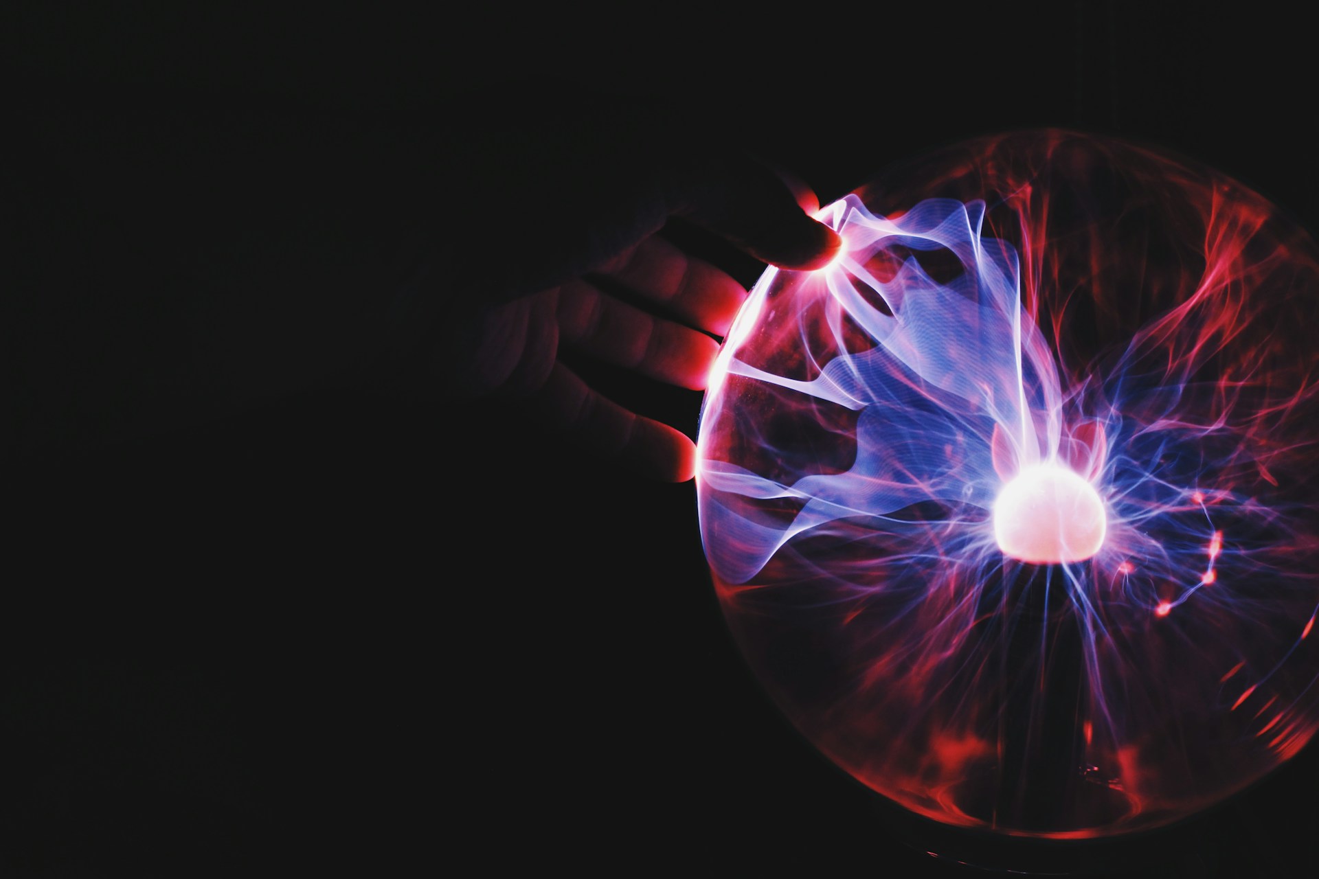 A hand touching the surface of a plasma globe, with colorful electric-like tendrils of blue and red light radiating towards the contact point, set against a dark background.